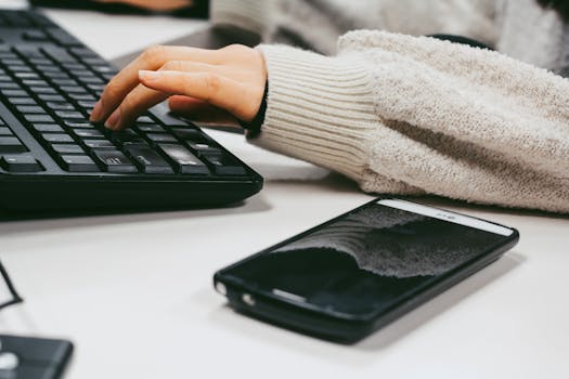 Close-up of a woman's hand typing on a keyboard with a smartphone placed on the desk.